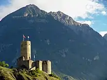 Photographie en couleurs d'un château-fort sur un éperon rocheux, donjon cylindrique et montagne à l'arrière-plan.