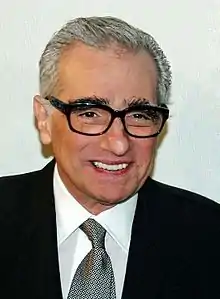 Photographie en gros plan d'un homme souriant aux cheveux gris qui porte des lunettes, habillé d'un costume élégant.
