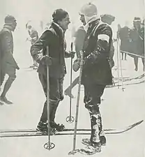 Photographie en noir et blanc d'une femme souriante et d'un homme moustachu à skis se faisant face.