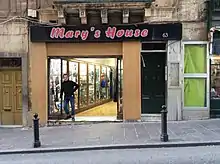 Photo d'un magasin à la devanture beige avec une inscription rouge sur fond noir en haut de celle-ci. Un homme se tient près de l'entrée, qui consiste en une série de vitrines et un renfoncement vers l'intérieur du magasin.