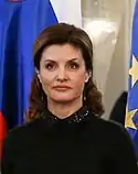 Gros plan sur une femme Blanche vêtue de noir se tenant face-caméra. Les cheveux de celle-ci sont bruns et ramenés vers l’arrière. En arrière plan, les drapeaux de l’Union Européenne et de la Russie sont visibles.