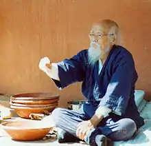 C'est une photographie en couleur de Fukuoka assis devant des récipients en céramique, il a un âge avancé.