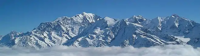  Les sommets culminants du Massif du Mont Blanc émergent d'une couche de nuages blancs dont on n'aperçoit qu'une mince bande en bas de la photo. Leur énorme masse enneigée se dresse donc au-dessus des nuages dans un ciel dégagé, parfaitement bleu.