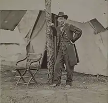 Photographie d'un homme en uniforme militaire prenant la pose en s'appuyant contre un arbre devant une large tente
