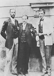 Un homme de petite taille en complet veston et portant moustache se tient debout, soutenu par deux infirmiers.