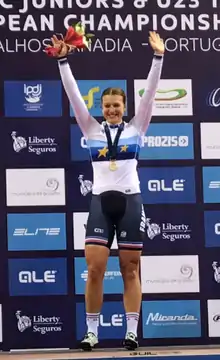 Une sportive portant un maillot de champion d'Europe lève les bras sur podium, médaille autour du cou.