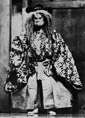 Acteur debout, figé, en tenue traditionnelle avec un masque massif et sévère et une large perruque.