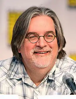 Photo en buste d'un homme au visage rond, portant des lunettes, une barbe blanche et des cheveux mi-longs, habillé en chemise à carreaux.