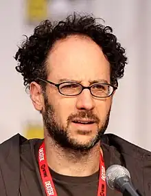 Photographie du buste d'un homme assis. Il a des cheveux bouclés, une paire de lunettes noires et un porte badge rouge.