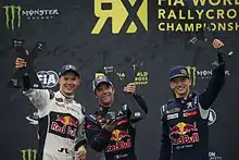 Trois hommes en combinaison de pilote automobile vus de face et soulevant chacun un trophée.