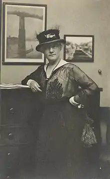 La photo en noir et blanc montre Mattie Edwards Hewitt, portrait de trois quarts de longueur, debout, face à l'avant, bras droit sur la commode,avec un chapeau.