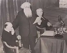 Photographie de trois hommes faisant une conférence. Francis Jammes est debout et lit un document.