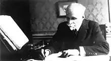 Photographie noir et blanc d'un homme écrivant, derrière son bureau.