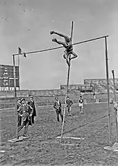 Photographie en noir et blanc d'un homme sautant à la perche dans un stade sous le regard d'hommes en costumes.