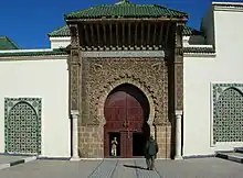 Photographie d'une tour brune sombre ouverte d'une porte avec un arc outrepassé, richement décoré et couvert d'un toit vert (oxyde de cuivre ou céramique). L'ensemble est flanqué de deux murs blancs percés d'ouvertures plus simples.