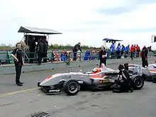 Photographue d'une monoplace de Formule 3 grise avec des teintes rouges, vue de profil, dans la voied es stands.