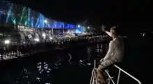 Photo de nuit. Assis sur le balcon avant de son bateau qui approche du quai, le navigateur salue la foule qui l'attend.