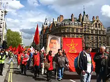 Photo couleur de manifestants défilant dans une rue sous un ciel bleu nuageux et portant des drapeaux rouges et un portrait de Staline.