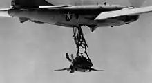 Photographie en noir et blanc d'un petit avion de chasse suspendu à un grand bombardier en vol, via un trapèze.