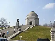 Bâtiment circulaire blanc surmonté d'un dôme au sommet d'une butte avec un escalier d'accès où se trouve une statue noire.