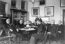 Groupe d'hommes en costumes assis autour d'une table dans un bureau richement décoré avec de nombreux portraits sur les murs.