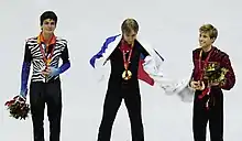 Photographie de trois patineurs en costume sur un podium, une médaille autour du cou, les deux à l'extérieur portant un bouquet de fleurs, l'un au centre entouré du drapeau russe.