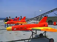 Trois drones rouges fuselés avec rampe sur pont de porte-avion. Les ailes delta et les nez sont jaunes (3 × 2,5 m env.).