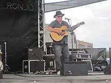 Photo d'un homme blanc debout sur une scène derrière un micro à pied en train de jouer de la guitare. Il porte un chapeau noir à large bord et des lunettes de soleil