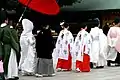 Photo couleur d'une procession de mariage dans l'enceinte d'un sanctuaire. Sept personnes sont visibles de dos, dont deux jeunes femmes, portant un haut blanc imprimé de motifs (branches d'arbre feuillues noires) et une sorte de jupe, longue et rouge.