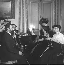 Photographie de 1910 représentant Mel Bonis dans son appartement parisien, dans un concert de musique de chambre. Mel Bonis se fait tourner les pages par sa belle-fille, Renée Fasquelle.