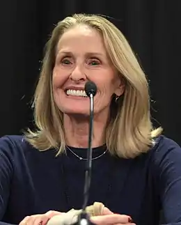 Photo en buste d'une femme âgée souriante avec des cheveux longs blonds et devant un micro.