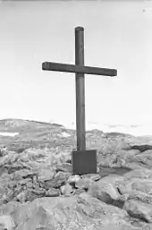 Photographie d'une grande croix en bois, posée verticalement au milieu de cailloux ; au bas de laquelle est fixée une plaque sur laquelle figurent des inscriptions.