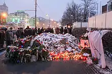 Plusieurs bougies et gerbes de fleurs sous la neige déposées au sol entourées de personnes.