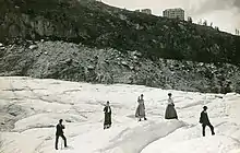 Photo en noir et blanc de touristes sur un glacier et, sur la crête en arrière-plan, trois bâtiments de taille inégale.