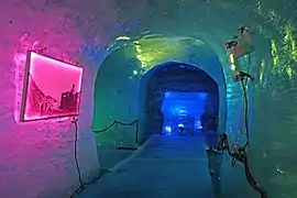 Intérieur d'une grotte de glace éclairée en rose et bleu avec un tableau sur la paroi et une sculpture creusée au fond.