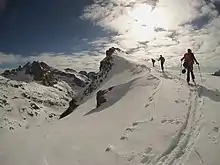 Trois skieurs en randonnée à l'approche d'un sommet, et d'autres sommets enneigés au second plan.