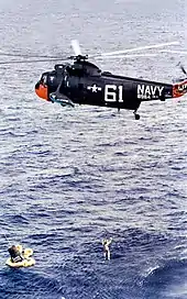 Un hélicoptère sort un homme en combinaison spatiale de l'eau avec un treuil. Un vaisseau spatial visible à la gauche de l'homme.