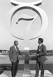 Photographie en noir et blanc de Grissom et Schirra à côté d'un monument représentant l'emblème du programme Mercury.