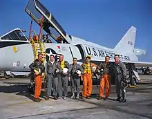 Les astronautes posent par ordre alphabétique devant un avion à réaction blanc à ailes delta. Ils tiennent leur casque de vol sous les bras. Les trois aviateurs de la marine portent des combinaisons de vol orange ; ceux de l'armée de l'air et de la marine portent du vert.