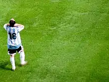 Photographie couleur. Homme revêtant la tenue de l’équipe d’Argentine de football se tient debout, de dos, sur une pelouse. Sur son maillot est marqué  « Messi » et « 18 ».