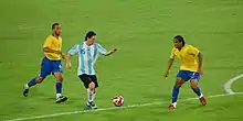 Photographie couleur. Au premier plan, Messi joue pour l’Argentine à l’entrée de la surface de réparation. Ses adversaires lui disputant la balle sont au nombre de deux et revêtent la tenue de l’équipe du Brésil de football.