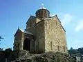 église en forme de croix au sol bâtie en grosses pierres de taille roses, grises et sable dont le clocher est surmonté par un croix orthodoxe devant un ciel bleu azur.