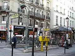 Photo en couleurs sur fond d'immeubles de rapport parisiens d'une bouche de métro flanquée derrière d'un kiosque à journaux et sur le côté de deux boîtes à lettres jaunes taguées