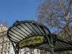 Photo en couleurs sur fond d'immeubles et de ciel bleu d'une petite toiture en verre avec marquise arrondie cerclées de fer forgé