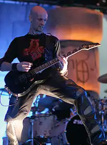 Photo en couleur d'un homme chauve en tee-shirt noir jouant de la guitare sur une scène