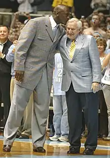 Michael Jordan en costume gris et cravate embrassant affectueusement le crâne de son ancien coach, Dean Smith, sur un parquet de basket-ball