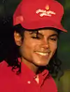 A headshot of a man wearing a red baseball cap and shirt. He has long black hair and is smiling towards the camera.