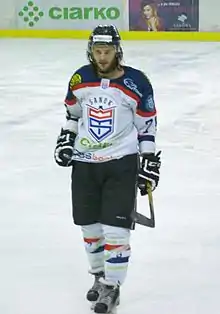 Photographie couleur d’un joueur de hockey sur glace, de face, en pied, portant une barbe de quelques jours et les cheveux dépassant du casque