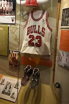 Tenue de match de Michael Jordan présentée dans une vitrine (de bas en haut) : une paire de chaussures noires de basket-ball, un maillot blanc siglé de rouge portant le numéro 23 des Bulls et une casque humoristique rouge siglé du logo des Bulls avec des cornes de taureau.