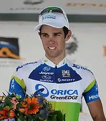 Photographie d'un homme portant une casquette et par dessus des lunettes de soleil et tenant dans sa main droite un bouquet de fleurs.